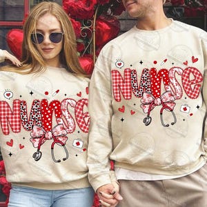 May include: Beige sweatshirts with the word "NURSE" in red and white patterned letters. Each sweatshirt features a bow, stethoscope, hearts, and medical-themed graphics. The sweatshirts are a matching set.