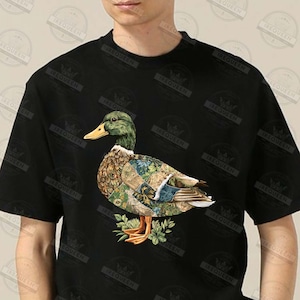 May include: Black t-shirt featuring a detailed illustration of a duck with a green head, yellow beak, and patterned body. The duck is standing on a bed of green leaves. The shirt has the word "IREQUEEN" printed on it.