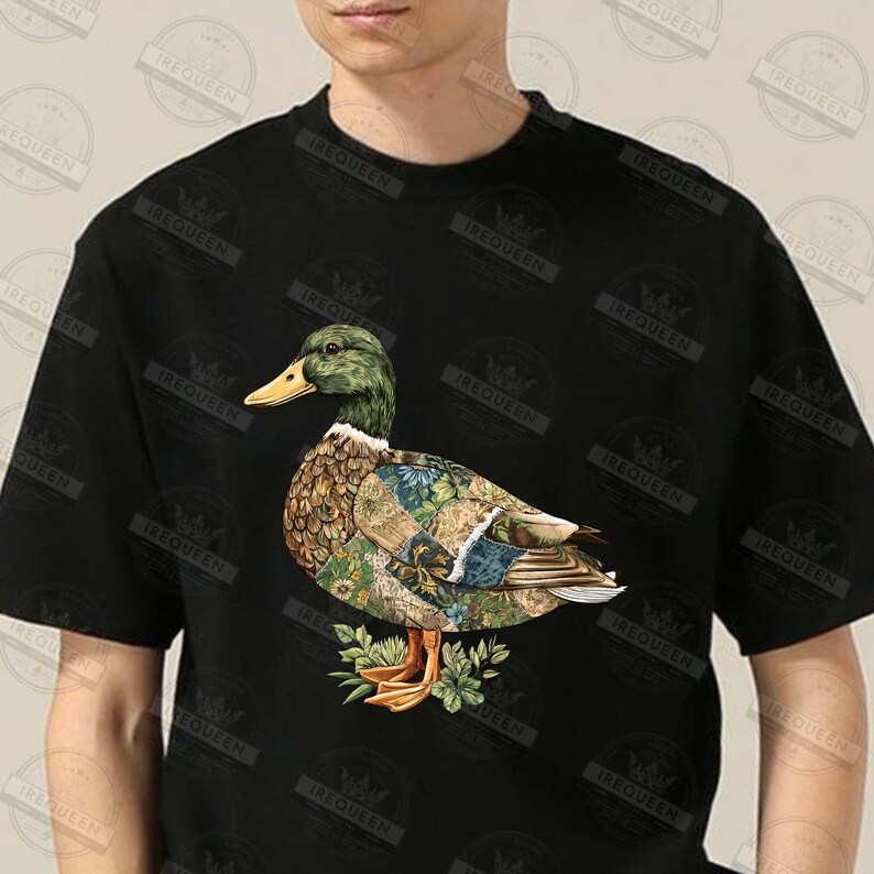 May include: Black t-shirt featuring a detailed illustration of a duck with a green head, yellow beak, and patterned body. The duck is standing on a bed of green leaves. The shirt has the word "IREQUEEN" printed on it.