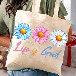 Pu&ograve; includere: Una borsa tote beige con tre illustrazioni di margherite colorate in blu, rosa e bianco, ciascuna con un centro giallo. La frase "Life is Good" &egrave; scritta in corsivo sotto i fiori. La borsa ha un design semplice con manici lunghi.