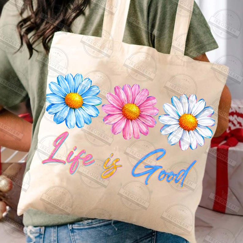 Pu&ograve; includere: Una borsa tote beige con tre illustrazioni di margherite colorate in blu, rosa e bianco, ciascuna con un centro giallo. La frase "Life is Good" &egrave; scritta in corsivo sotto i fiori. La borsa ha un design semplice con manici lunghi.