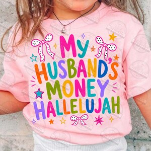 Pu&ograve; includere: Una t-shirt rosa chiaro con il testo colorato My Husband's Money Hallelujah. Il design include fiocchi bianchi a pois neri, insieme a varie piccole stelle nelle tonalit&agrave; del blu, arancione, giallo e rosa sparse sulla parte anteriore.