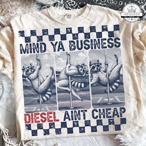 Puede incluir: Sudadera color crema con un gráfico que presenta a tres mapaches bailando en barra. El texto "MIND YA BUSINESS" está encima de los mapaches y "DIESEL AINT CHEAP" debajo. El diseño incluye un patrón de cuadros.