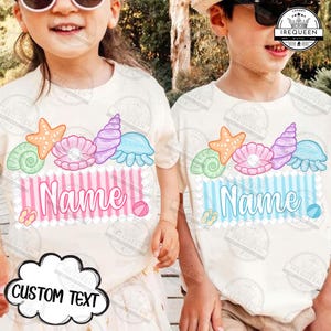 May include: Two children wearing off-white t-shirts with a seashell design. The shirts feature a starfish, shells, and the word "Name" in a striped box. The design includes a pink and blue color scheme, with a beach theme.