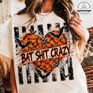 May include: A cream-colored t-shirt featuring a graphic of an orange heart filled with a black spiderweb pattern. The design includes black bats, spiders, and vertical stripes, with the text BAT SHIT CRAZY printed in a distressed black font.