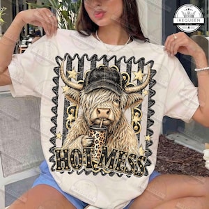 Può includere: Una t-shirt color crema con la grafica di una mucca Highland che indossa un berretto da baseball nero e tiene in mano una tazza con stampa leopardata. Il design è incorniciato da un bordo a righe bianche e nere con stelle dorate e la scritta HOT MESS.