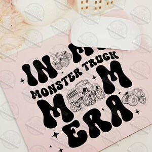 May include: A pink mouse pad with black text that reads "In the Monster Truck Mom Era". The text is in a retro font style. There are two black and white illustrations of monster trucks on the mouse pad.