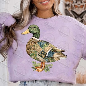 May include: A purple sweatshirt featuring a detailed illustration of a duck. The duck has a green head, yellow beak, and a body adorned with floral and patterned designs. The duck is standing on a bed of green leaves.