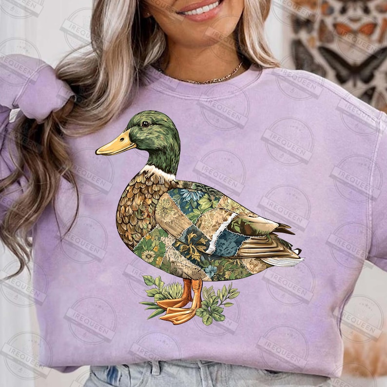 May include: A purple sweatshirt featuring a detailed illustration of a duck. The duck has a green head, yellow beak, and a body adorned with floral and patterned designs. The duck is standing on a bed of green leaves.