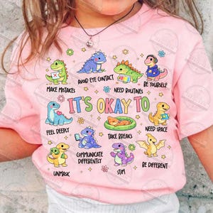 May include: A pink t-shirt with cartoon dinosaurs and phrases such as "It's Okay To" and "Need Space." The design includes various colours and is suitable for children. The t-shirt has a round neck and short sleeves.