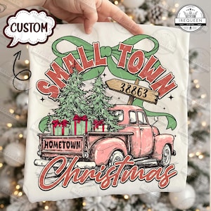 May include: A white t-shirt featuring a pink vintage truck carrying Christmas trees and gifts. The text reads "Small Town Christmas" with a hometown sign and the number 38863. A green bow and stars complete the design.