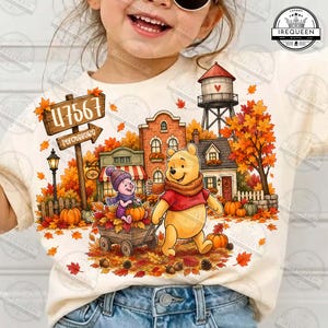 May include: A cream-colored t-shirt featuring a colorful autumn scene with Winnie the Pooh and Piglet. The graphic includes a wooden sign reading 47567 Petersburg, a water tower, a coffee shop, and pumpkins amidst orange and red fall foliage.