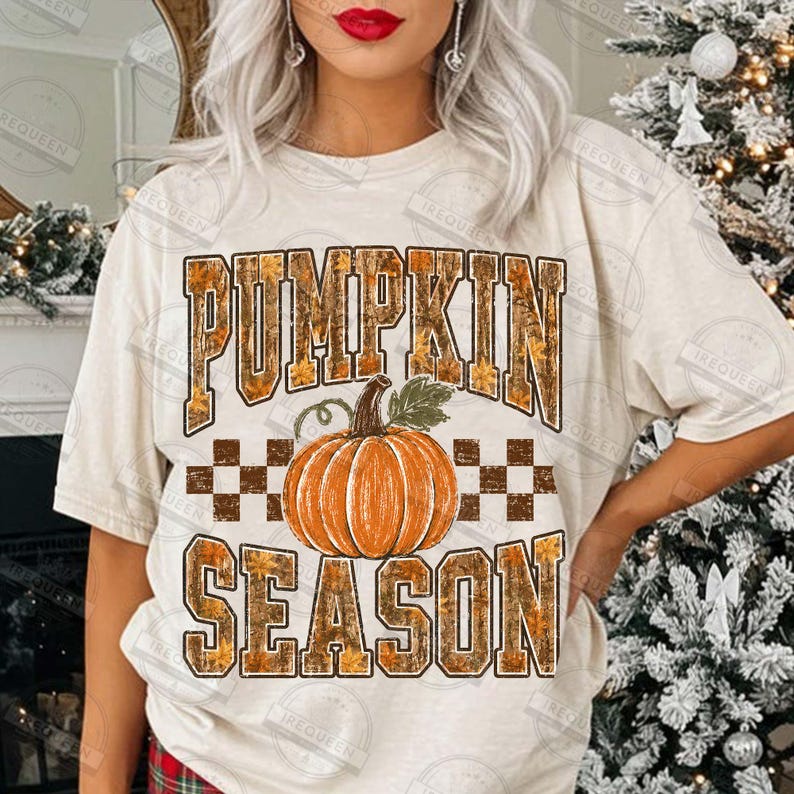 May include: A cream-colored short-sleeve t-shirt featuring a graphic print with the text PUMPKIN SEASON in a brown and orange leaf-patterned font. A central orange pumpkin illustration sits above the word SEASON with a checkered pattern.