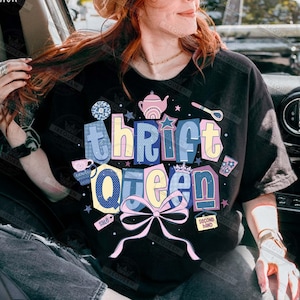 May include: Black t-shirt with the words "Thrift Queen" in a colorful, layered design. The design includes a pink bow, stars, and illustrations of a teapot, teacup, and spoon. The words "sale" and "second hand" are also included.