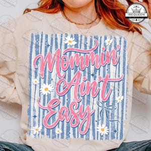 May include: A light-colored sweatshirt with the phrase "Mommin' Ain't Easy" in pink script. The design features a blue and white striped background, white daisies, and small blue stars. A bow graphic is also included.