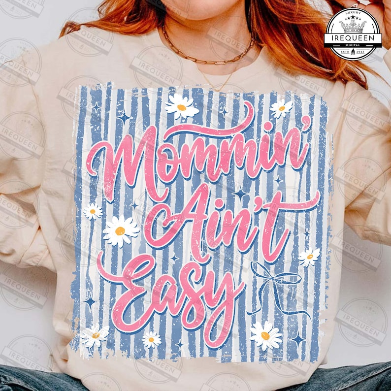 May include: A light-colored sweatshirt with the phrase "Mommin' Ain't Easy" in pink script. The design features a blue and white striped background, white daisies, and small blue stars. A bow graphic is also included.