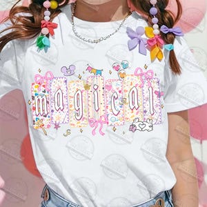 May include: White t-shirt with the word "magical" in a colorful, whimsical design. The letters are in a decorative font, each with a unique pattern and adorned with bows, stars, and cartoon elements. A playful and fun graphic tee.