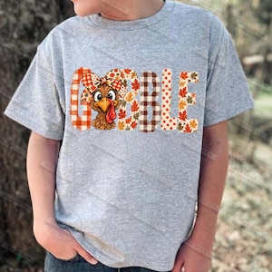 May include: A heather gray short-sleeve t-shirt featuring the word GOBBLE printed in large letters. The letters are filled with patterns of orange plaid, brown and white buffalo check, and autumn leaves. A cartoon turkey with a plaid bow is included.