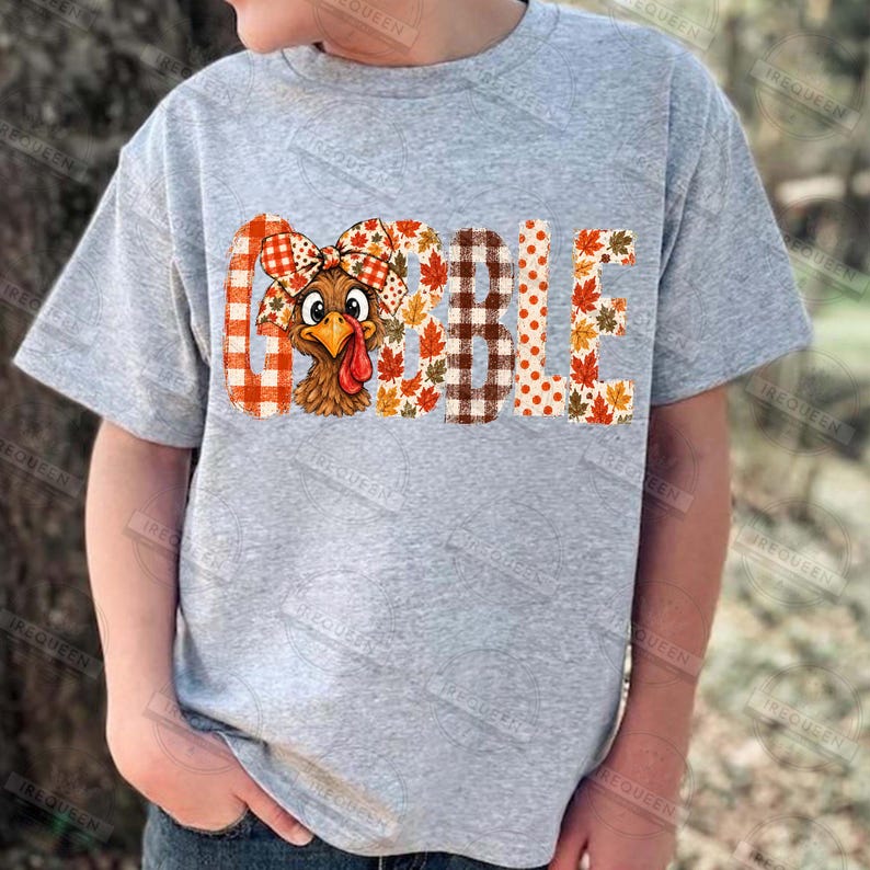 May include: A heather gray short-sleeve t-shirt featuring the word GOBBLE printed in large letters. The letters are filled with patterns of orange plaid, brown and white buffalo check, and autumn leaves. A cartoon turkey with a plaid bow is included.