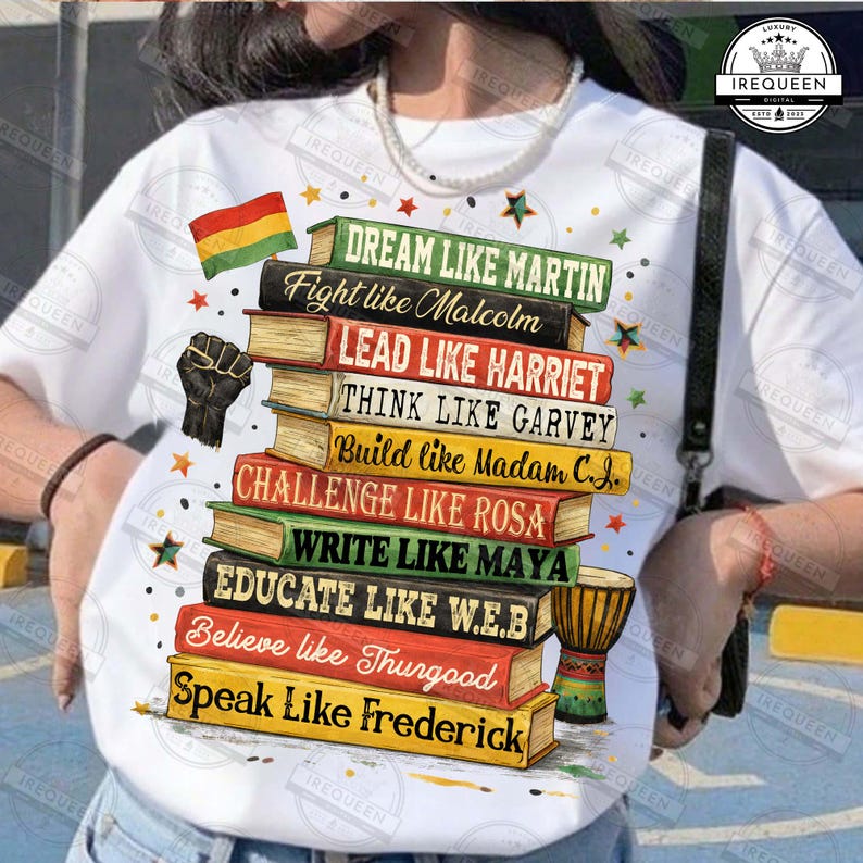 May include: White t-shirt featuring a stack of books with inspirational phrases like "Dream Like Martin" and "Speak Like Frederick." The design includes a black power fist, a flag, stars, and a small drum. The shirt is a statement piece.