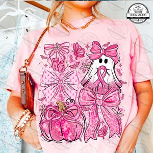 May include: Pink t-shirt featuring a collection of pink bows, a ghost, a pumpkin, and boxing gloves. The design includes pink ribbons, butterflies, and bats. The shirt has the text "IREQUEEN" on it.