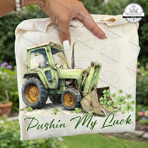 May include: A light beige tote bag featuring a green tractor illustration with gold wheels and a front loader. The text "Pushin My Luck" is written in green script below the tractor, with shamrocks scattered around.