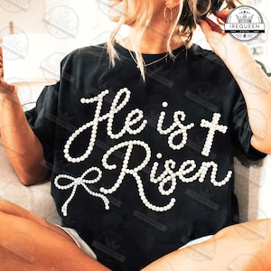 May include: Black t-shirt with the words "He is Risen" in white, pearl-like lettering. The design includes a cross and a bow, creating a religious theme. The shirt is a casual wear item.