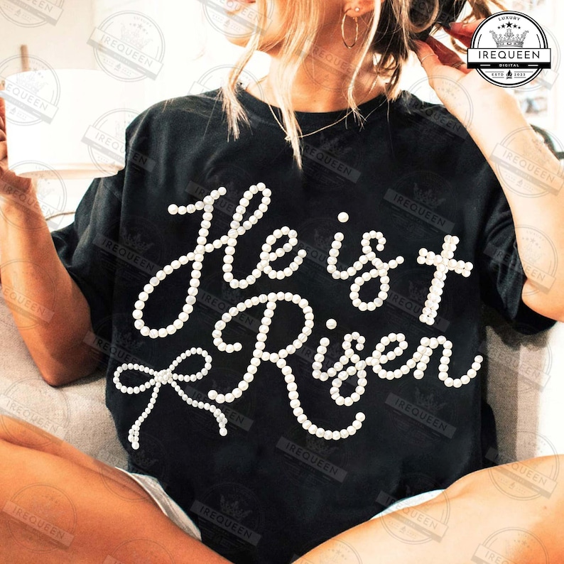 May include: Black t-shirt with the words "He is Risen" in white, pearl-like lettering. The design includes a cross and a bow, creating a religious theme. The shirt is a casual wear item.