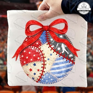 Puede incluir: Camiseta blanca con un diseño de béisbol con parches rojos, blancos y azules. Un lazo rojo adorna la parte superior. El diseño de béisbol incluye estrellas, rayas y equipamiento de béisbol.