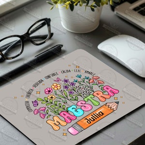 May include: A gray mousepad with a colorful illustration of the word "MAESTRA" with a pencil and flowers. The pencil is labeled "Jullia". The words "COMPASIVA CARINOSA DEDICADA CONFIABLE CALIDA LEAL AMABLE" are written around the flowers.