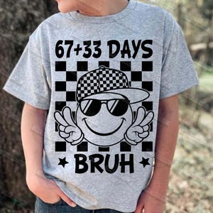 Pu&ograve; includere: T-shirt grigio chiaro con una grafica di una faccina sorridente con occhiali da sole e un cappello a scacchi, con segni di pace. Il testo recita "67+33 DAYS BRUH" in nero. Lo sfondo &egrave; un motivo a scacchi.