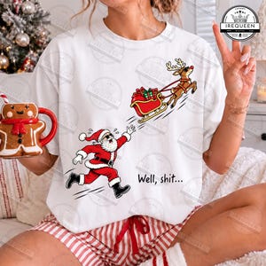 May include: A white short-sleeve t-shirt featuring a graphic of Santa Claus running after a reindeer pulling a sleigh filled with gifts. The text "Well, shit..." is printed below the graphic. The shirt is paired with red and white striped shorts.