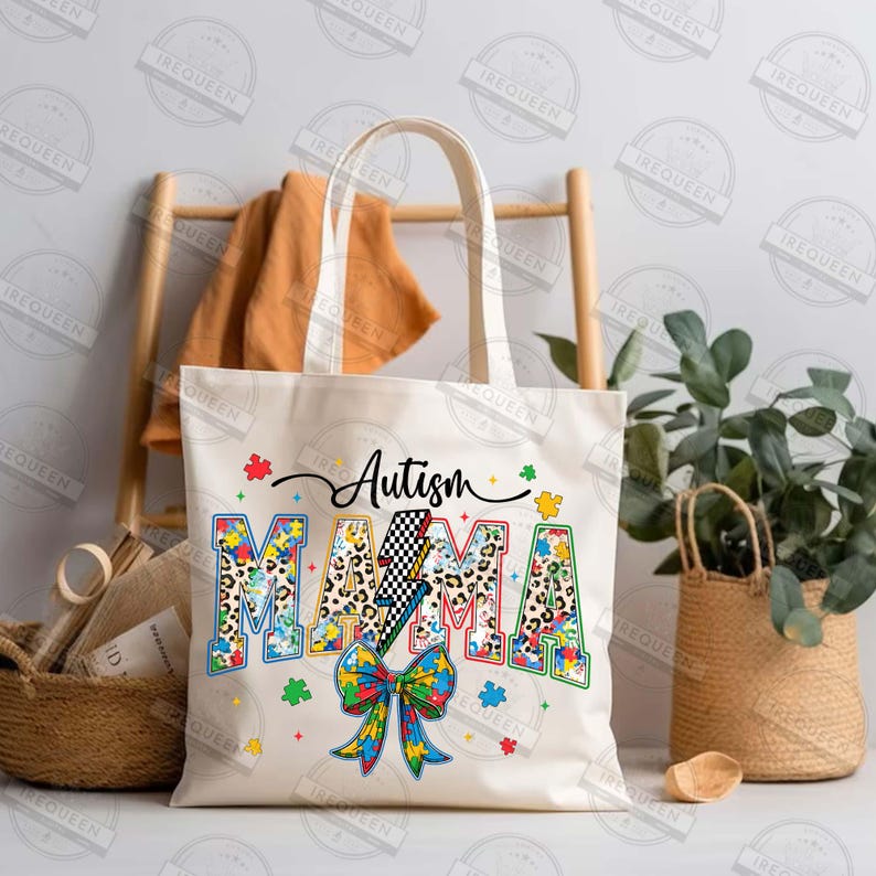 May include: A white canvas tote bag with the text "Autism Mama" in colorful letters. The letters are decorated with a leopard print pattern and puzzle pieces. A blue and yellow ribbon with a puzzle piece in the center is below the text.