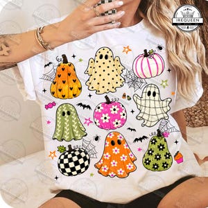 Puede incluir: Una camiseta blanca con un estampado gráfico colorido con temática de Halloween. El diseño incluye varios fantasmas y calabazas decorados con patrones como lunares, flores y cuadros, intercalados con pequeñas telarañas, murciélagos y estrellas.
