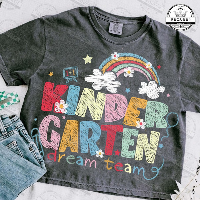 May include: Dark gray t-shirt with the words "KINDERGARTEN dream team" in colorful, sketched letters. A rainbow, clouds, and stars decorate the shirt. A pair of blue jeans is visible in the lower left corner.