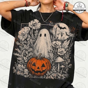 May include: A black t-shirt featuring a graphic print of a central white ghost surrounded by orange pumpkins, skulls, and dark foliage. The design includes a bat silhouette against a full moon, with intricate botanical details and a spooky aesthetic.