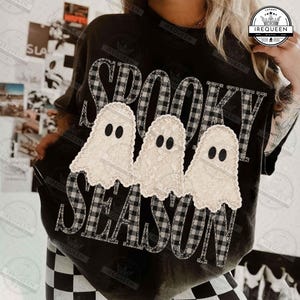 Può includere: Una t-shirt nera con la scritta SPOOKY SEASON in un motivo a quadri bianchi e neri. Tre figure di fantasmi color bianco sporco con una trama in pizzo sono posizionate al centro del testo. Il design è stampato sulla parte anteriore.