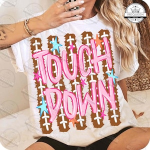 May include: A white short-sleeve t-shirt featuring a graphic print of brown football-style laces arranged in vertical stripes. The text TOUCH DOWN is printed in large, bright pink bubble letters, accented by small light blue and orange stars.