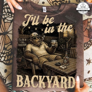 May include: A brown t-shirt featuring a graphic of a Bigfoot character wearing sunglasses and a cap, sitting in a backyard chair while holding a glass of whiskey and a cigar. The text reads I'll be in the BACKYARD. A bottle of whiskey sits on a table.