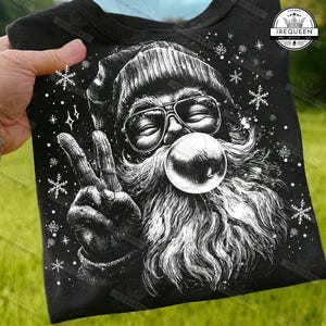 May include: Black t-shirt featuring a graphic of Santa Claus with sunglasses blowing a bubble. Santa is giving the peace sign. White snowflakes and stars are scattered around the image. The shirt is held by a hand.