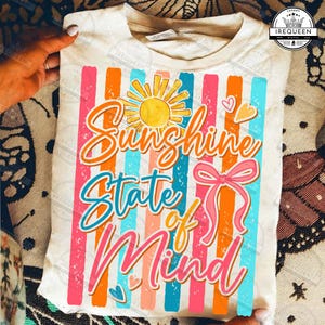 May include: Cream-colored t-shirt with the text "Sunshine State of Mind" in a colorful design. The design includes a yellow sun, pink bow, and vertical stripes in orange, pink, and blue.