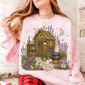 May include: A light pink sweatshirt featuring a graphic illustration of a wooden cottage surrounded by lavender plants, a stone path, a black cauldron with purple liquid, and a glass bottle labeled Witches Brew, accented by stars and a crescent moon.