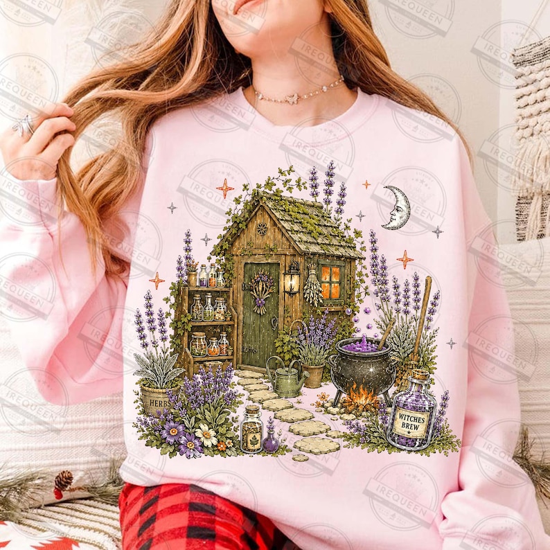 May include: A light pink sweatshirt featuring a graphic illustration of a wooden cottage surrounded by lavender plants, a stone path, a black cauldron with purple liquid, and a glass bottle labeled Witches Brew, accented by stars and a crescent moon.