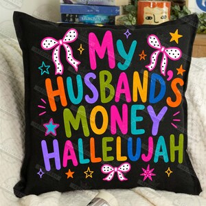 Pu&ograve; includere: Un cuscino quadrato nero con il testo colorato My Husband's Money Hallelujah in un carattere giocoso. Il design presenta lettere rosa brillante, arancioni, blu, verdi e viola, accentuate da fiocchi bianchi a pois e piccole stelle decorative.