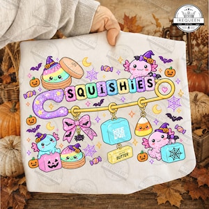 May include: A white t-shirt featuring a colorful graphic of a large safety pin with the text SQUISHIES. The design includes cute axolotls, pumpkins, bats, candy, a pink bow, a blue cube labeled NEEDOH, and a yellow block labeled SALTED BUTTER.