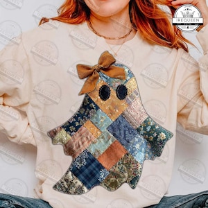 May include: A cream-colored sweatshirt featuring a patchwork ghost design. The ghost is made of various patterned fabrics in shades of blue, orange, and brown, with a brown bow at the top. The ghost has two black oval eyes.