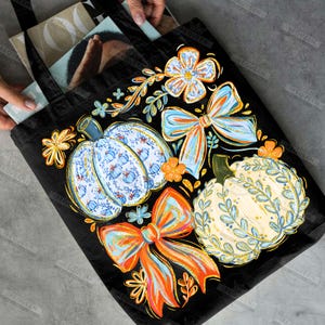 May include: Black tote bag featuring a colorful autumn design. The design includes pumpkins, bows, and floral elements in shades of blue, orange, and yellow. The tote bag is filled with books and magazines.