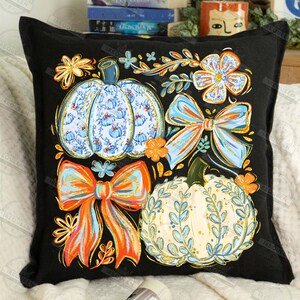 May include: Black square throw pillow featuring a colorful autumn design. The design includes two pumpkins, two bows, and floral accents in shades of blue, orange, and yellow. The pumpkins have intricate patterns, and the bows have a gradient effect.