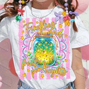 May include: White t-shirt featuring a colorful graphic of a mason jar filled with fireflies, with the text "Fireflies and front porch nights." The design includes pink stripes, stars, and floral elements.