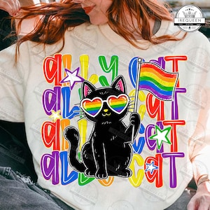 May include: A cream-colored t-shirt featuring a black cat wearing heart-shaped rainbow glasses and holding a rainbow flag. The background text reads "ALLY CAT" in repeating, colorful, bold letters, accented by small, multi-colored stars.